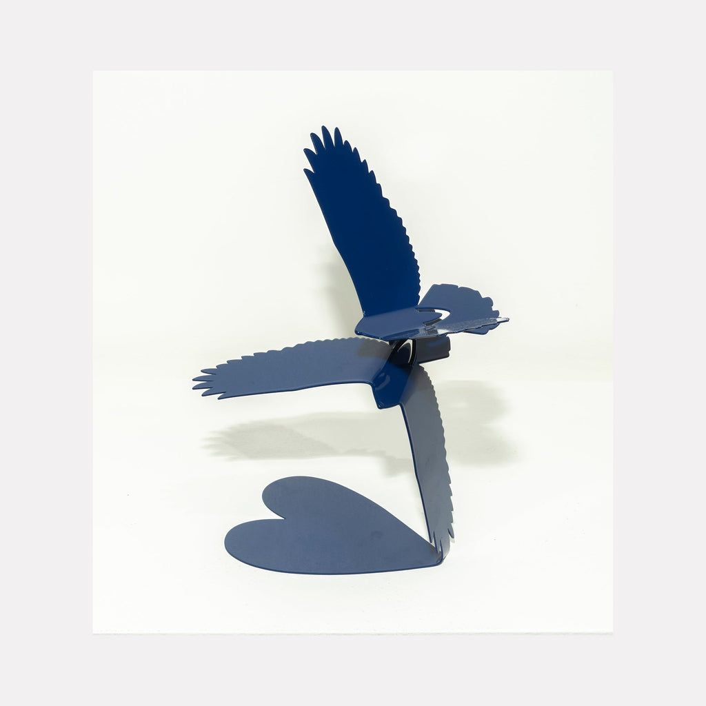 The artwork 'Love Birds Deep Blue' by Nicki Adani features a sculptural steel bird in flight, powder-coated in deep navy blue. The contemporary piece shows graceful wing spans with detailed feather cutouts, mounted on a heart-shaped base, creating a striking silhouette against white background. 12x12 inches metal art sculpture.