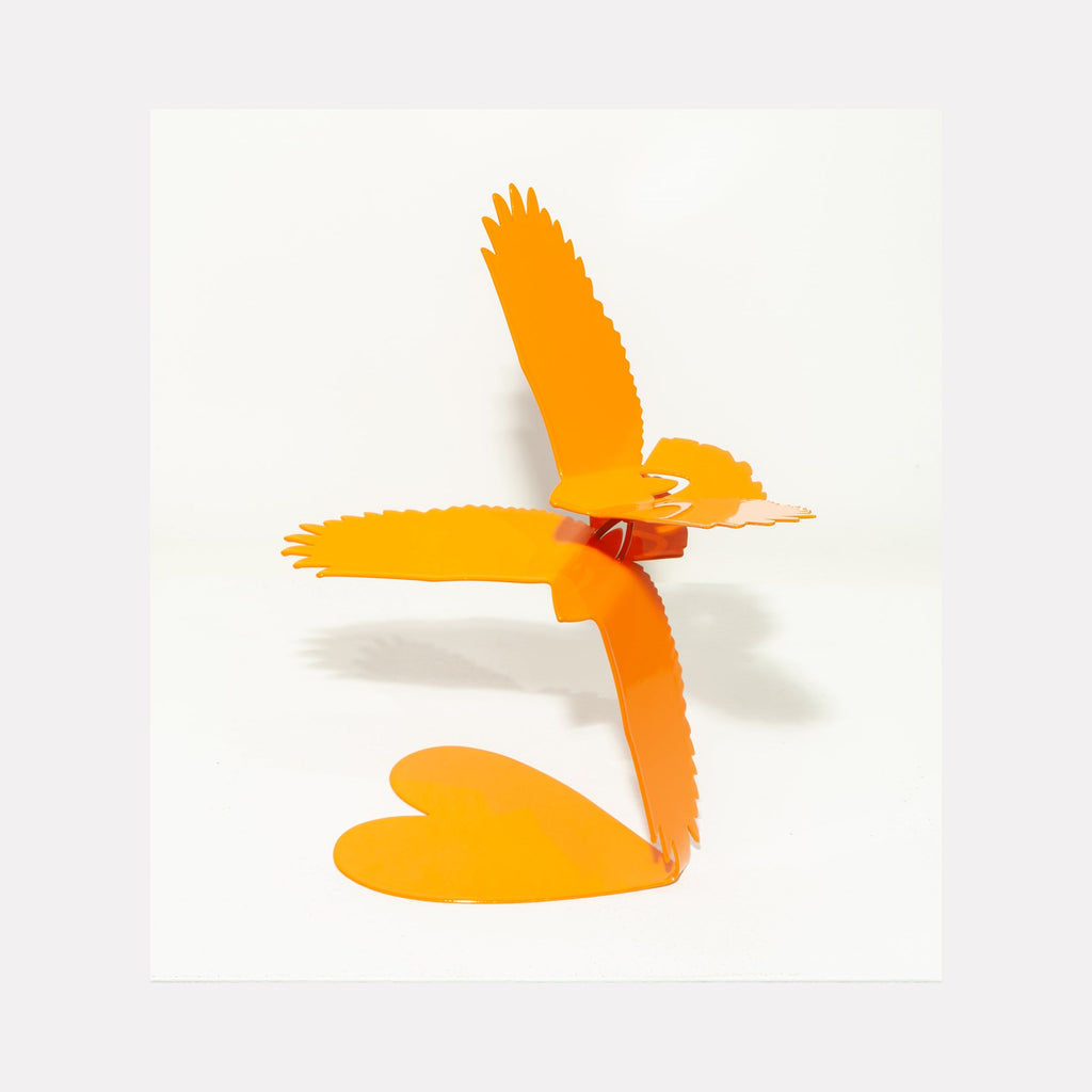 The artwork 'Love Birds Orange' by Nicki Adani features a vibrant orange steel sculpture depicting a stylized bird in flight with a heart-shaped base. The contemporary minimalist design showcases powder-coated steel craftwork with sleek lines and dynamic movement, measuring 12x12 inches. The striking silhouette casts gentle shadows on white background.