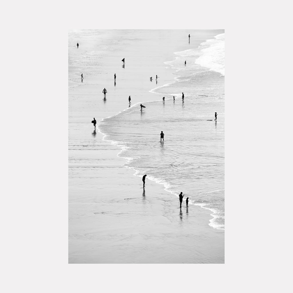 The artwork 'People on Beach' by Neil Shapiro captures silhouettes scattered along a serene shoreline in black and white photography. Figures appear as small dark shapes against light sand and gentle waves, creating a minimalist composition with strong visual contrast. Printed on textured Hahnemule paper, the aerial perspective emphasizes the peaceful solitude of beachgoers along the water's edge.