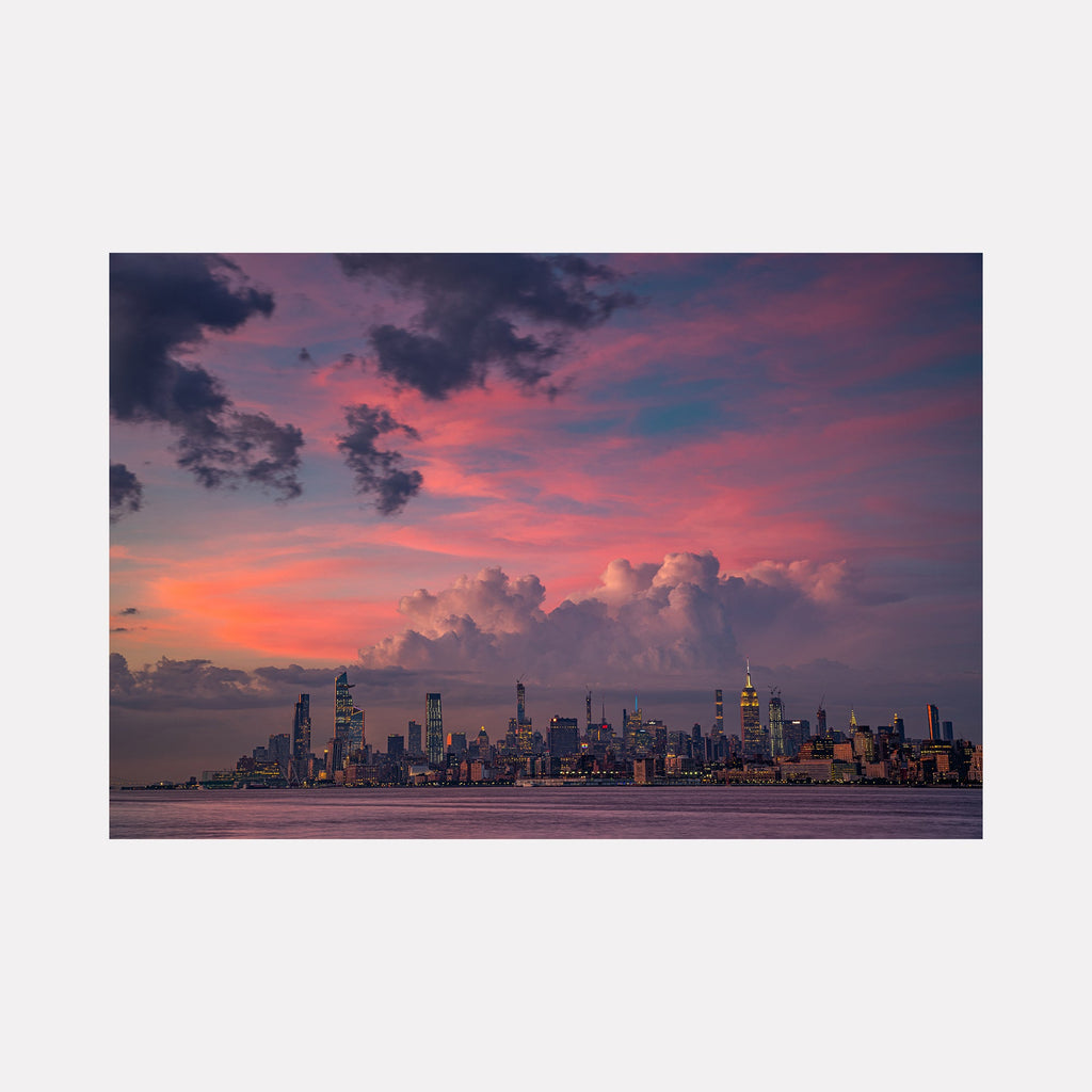 The artwork 'Pink Apple' by Dennis Maida captures New York City's iconic skyline at dusk, featuring dramatic pink and purple clouds illuminating the sky. Towering skyscrapers silhouetted against a vibrant sunset create a stunning urban panorama, with the Empire State Building and illuminated windows punctuating the cityscape reflected in calm harbor waters. Color photography. 16x25 inches.