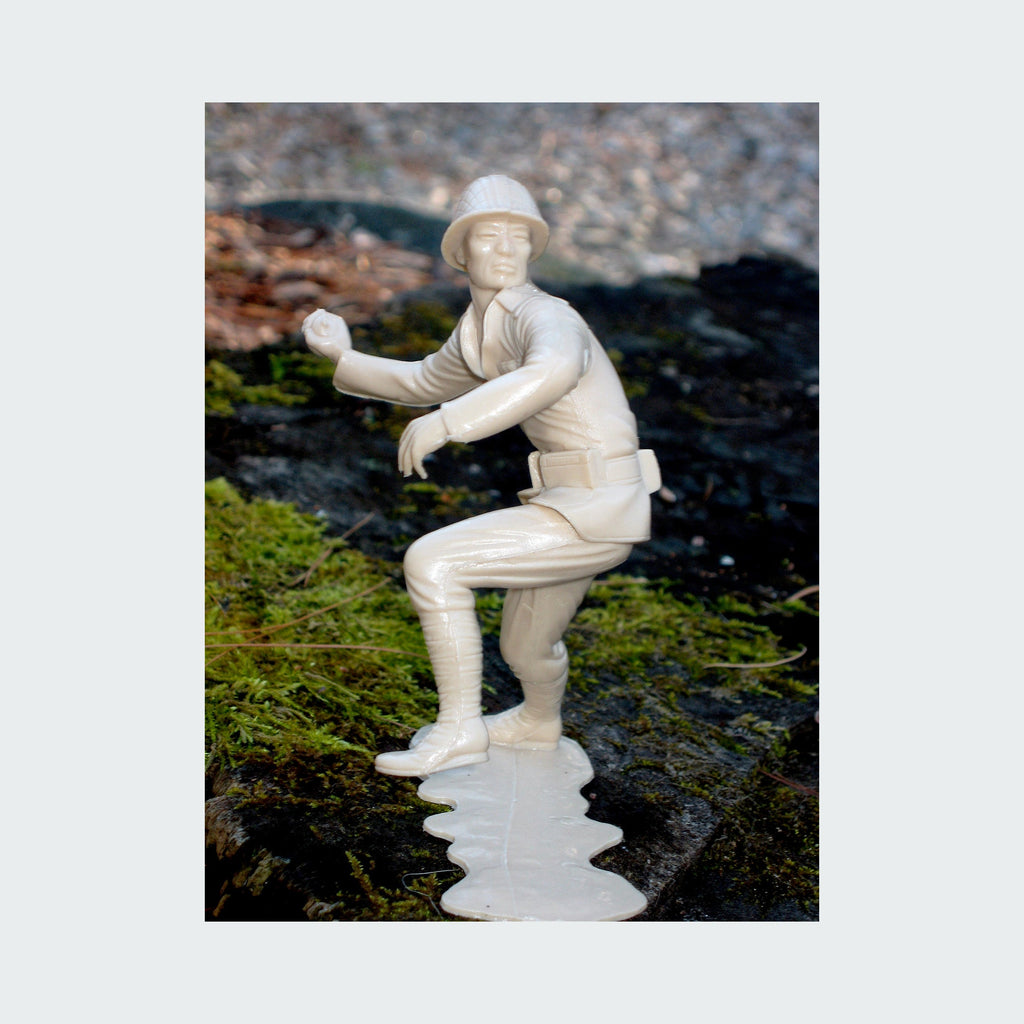 The artwork 'Plastic Men Series: Thrower' by Margaret Roleke features a monochromatic white plastic figurine in dynamic throwing pose, captured against a mossy rock backdrop. The sculpture shows meticulous detail in military-style uniform with helmet, displaying crisp lines and smooth texture characteristic of plastic molding.