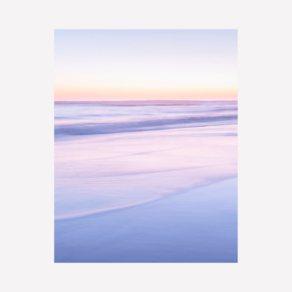 Portrait du point du jour #2 by Fabienne Sowa-Dobkowski captures a serene coastal sunrise in soft pastel hues. The photograph features gentle waves washing onto a beach, creating smooth horizontal lines in lavender, pink, and powder blue tones. The dreamy composition shows a misty morning atmosphere with seamless gradients from dawn sky to reflective water surface.