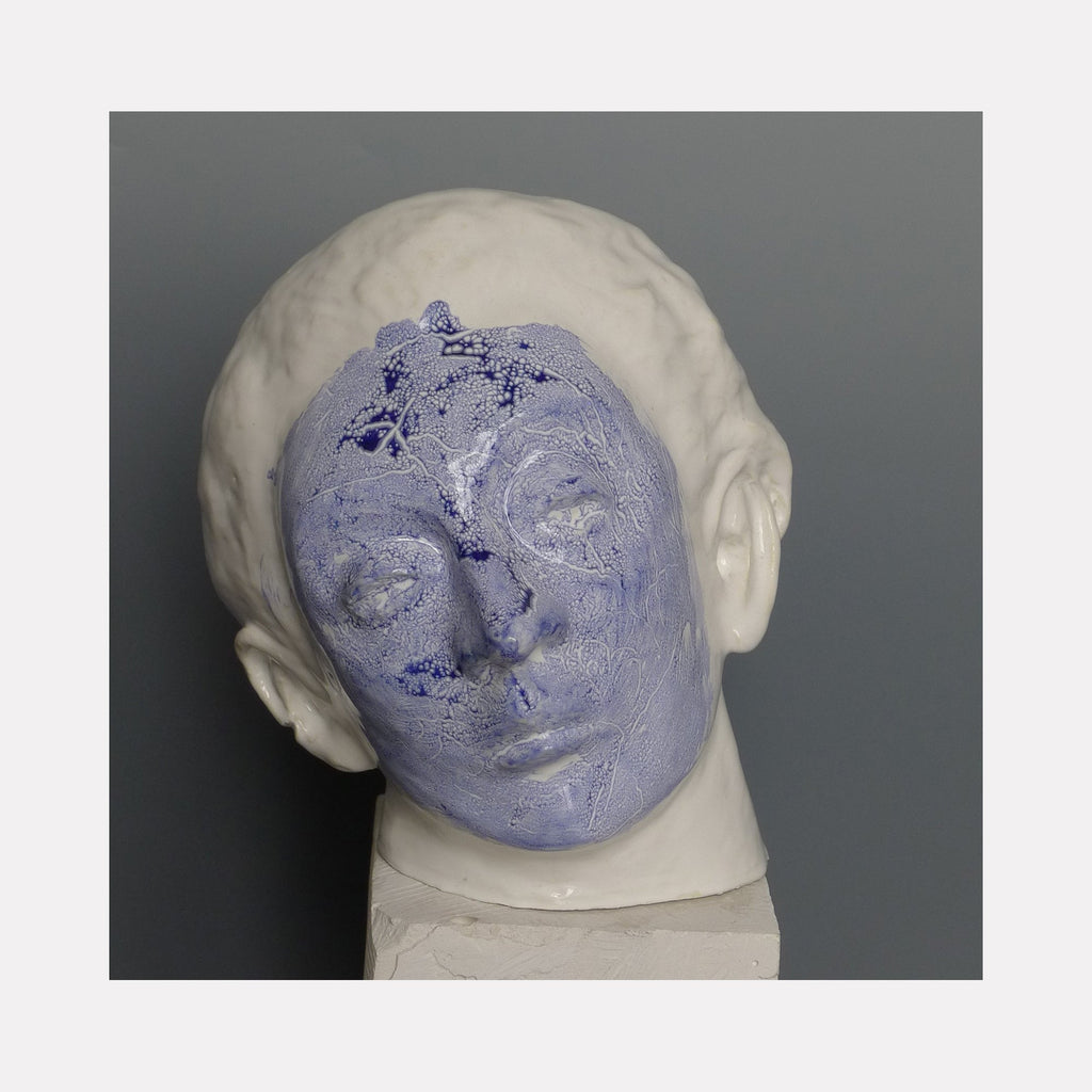 The artwork 'Prince J. Awakening from a Dream' features a striking ceramic head sculpture with a dual-toned finish - a lavender-blue textured face contrasting with smooth white glazed hair. The sculptural piece shows serene, dreamy facial features in a contemporary artistic style. Glazed ceramic artwork by Mark LaRiviere.