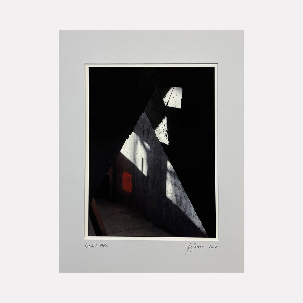 The artwork 'Rabbit Hole' by Cora Jane Glasser features dramatic contrasts of light and shadow in a minimalist composition. Sharp angular beams of white light pierce through darkness, creating geometric patterns against a black background. A subtle hint of red adds depth to the shadowed areas. Digital photograph printed on velvet archival paper captures architectural abstract qualities.