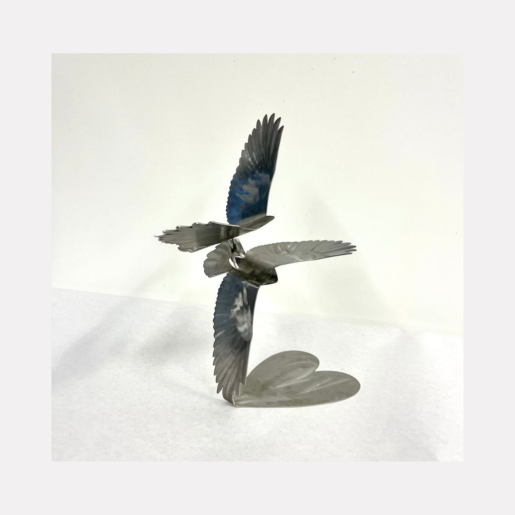 The artwork 'Love Birds Cloud' by Nicki Adani features two intricately detailed steel birds in flight, rendered in powder-coated metal with subtle blue accents on their wings. The sculpture captures the graceful movement of birds soaring together, creating a heart-shaped shadow beneath. The 12-inch contemporary metal sculpture combines modern minimalism with natural motion. By Nicki Adani.