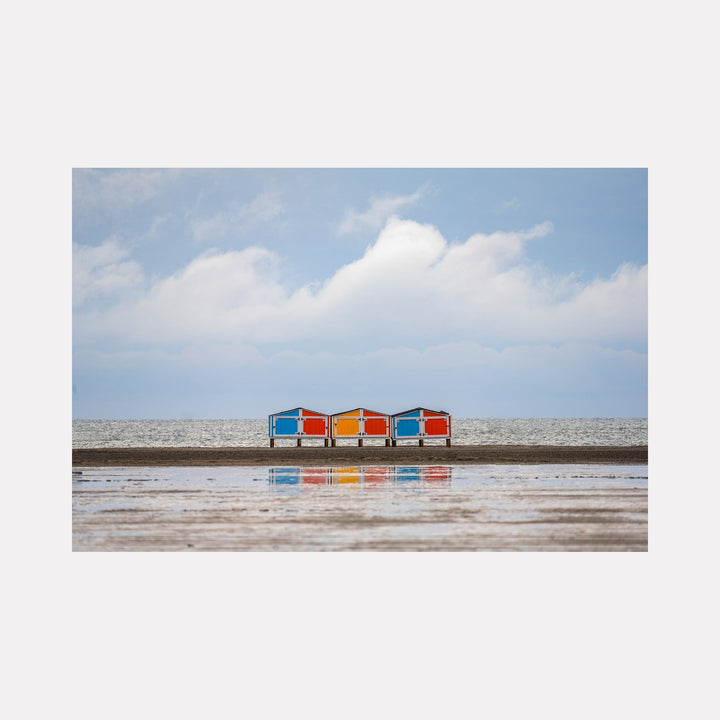 The artwork 'Sand and Sky' by Dennis Maida captures a serene beachscape featuring four colorful beach chairs - alternating blue and orange - lined up along a pristine shoreline. The chairs cast perfect reflections on wet sand beneath a soft blue sky dotted with wispy white clouds, creating a minimalist coastal composition at 16x20 inches. Photography by Dennis Maida.