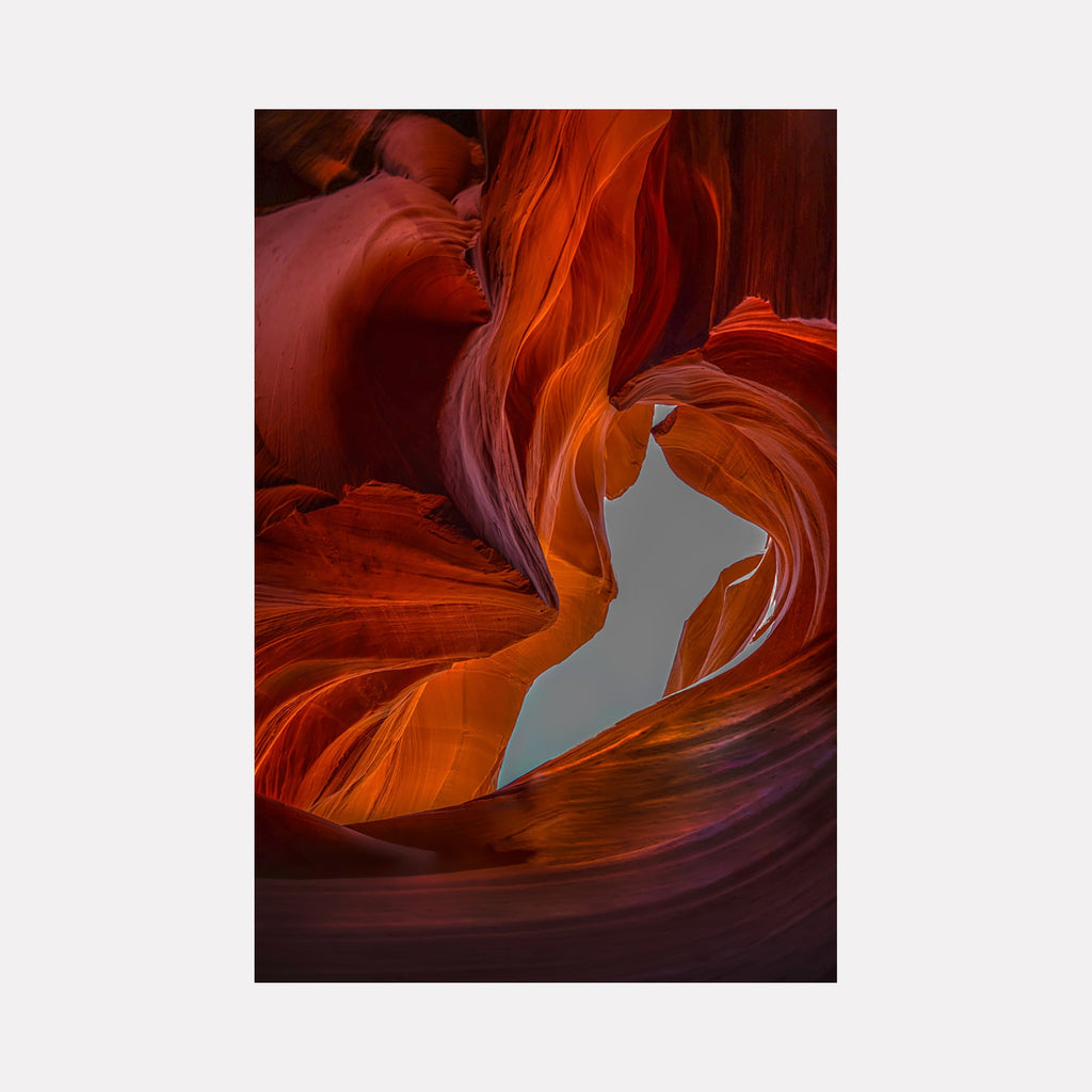 Slot Canyon In Color by Neil Shapiro captures dramatic red sandstone formations in Antelope Canyon, featuring flowing curves and smooth wave-like patterns illuminated by natural light. Rich orange and deep burgundy hues create striking contrasts while a patch of blue sky peeks through the curved canyon walls, printed on metal for enhanced vibrancy.