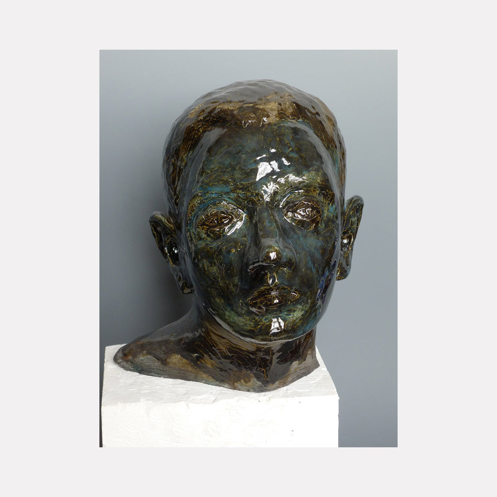 The artwork 'The Dark Prince' by Mark LaRiviere features a glazed ceramic head sculpture with iridescent dark patina, displaying a striking interplay of metallic bronze and blue-green tones. The sculptural bust exhibits smooth, polished surfaces with dramatic light reflections, creating a mysterious and ethereal effect. Mounted on a white pedestal, this 9x8x8 inch contemporary ceramic piece by Mark LaRiviere.