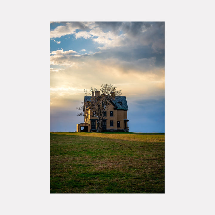 The artwork 'The House Where No One Lives' by Dennis Maida captures a solitary Victorian-style house standing against a dramatic sunset sky. The weathered yellow house with dark roof features bare trees alongside, set on an empty green field. Golden sunlight pierces through dramatic clouds, creating an ethereal atmosphere that emphasizes the building's abandoned solitude.