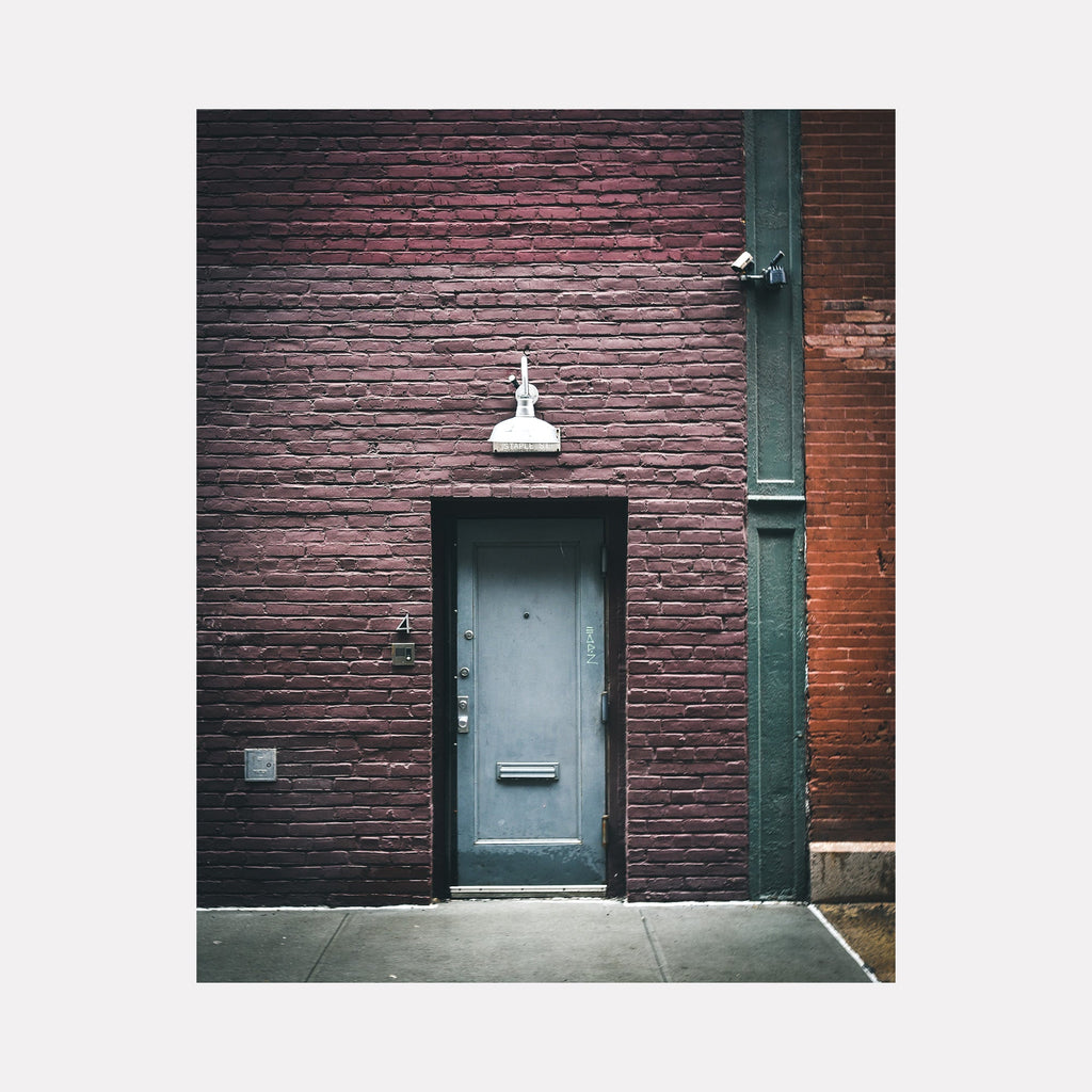 The artwork 'Thresholds Untold' by Dennis Maida captures a minimalist urban scene featuring a weathered teal door set against a textured burgundy brick wall. A vintage white industrial lamp hangs above the door, creating stark contrast. The composition emphasizes geometric lines and industrial architecture in modern photography, measuring 20x16 inches.