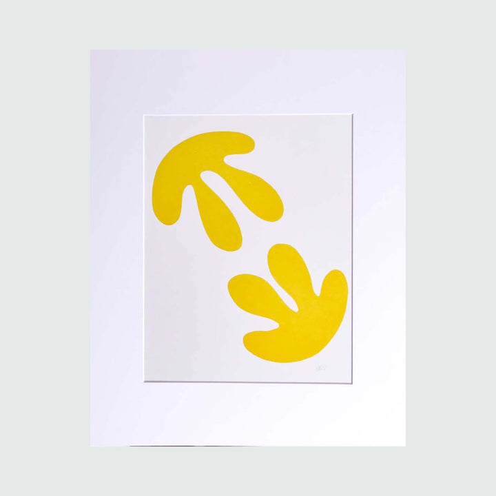 The artwork 'The Morning Meditation Series 1' by Shadia Sabagh features two abstract yellow organic shapes against a crisp white background. The minimalist freehand drawing showcases flowing, leaf-like forms arranged in a balanced composition, reminiscent of Matisse's cut-out style. Limited edition print, 20x16 inches.