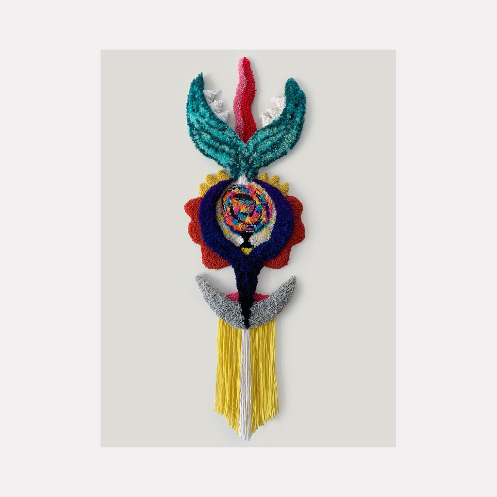 The artwork 'Carnivora' by Ana Maria Farina features a striking fiber sculpture combining turquoise crescent shapes, vibrant red petals, and a central spiral pattern in mixed colors. Yellow tassels cascade from silver wings at the base, creating a vertical totem-like composition on linen. Crafted from upcycled fibers, the piece measures 53.5 inches tall by 17 inches wide.