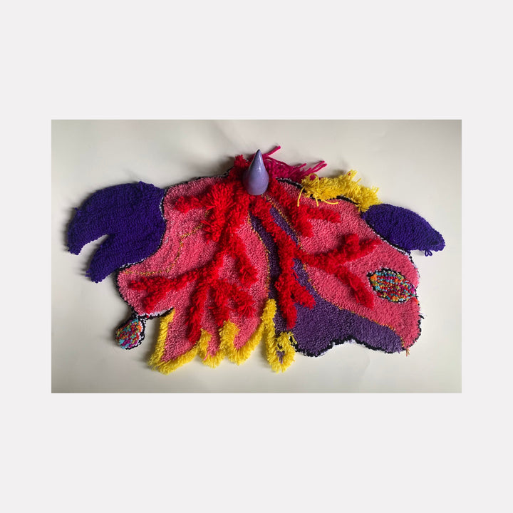 The artwork 'Horny #1' features a vibrant abstract textile composition with flowing forms in bright red, deep purple, and golden yellow upcycled fibers on linen. A small lavender ceramic element protrudes from the textured surface, creating a dynamic three-dimensional effect. The piece demonstrates organic, fluid shapes reminiscent of natural forms. By Ana Maria Farina.