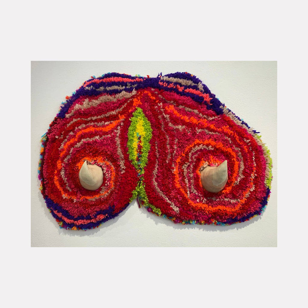 The artwork 'Horny #2' by Ana Maria Farina features a symmetrical abstract design with vibrant red swirling patterns reminiscent of butterfly wings. Two ceramic spheres are embedded within circular motifs, accented by bright neon green, deep purple, and orange upcycled fibers on linen. The textural piece creates a dynamic interplay of colors and materials.