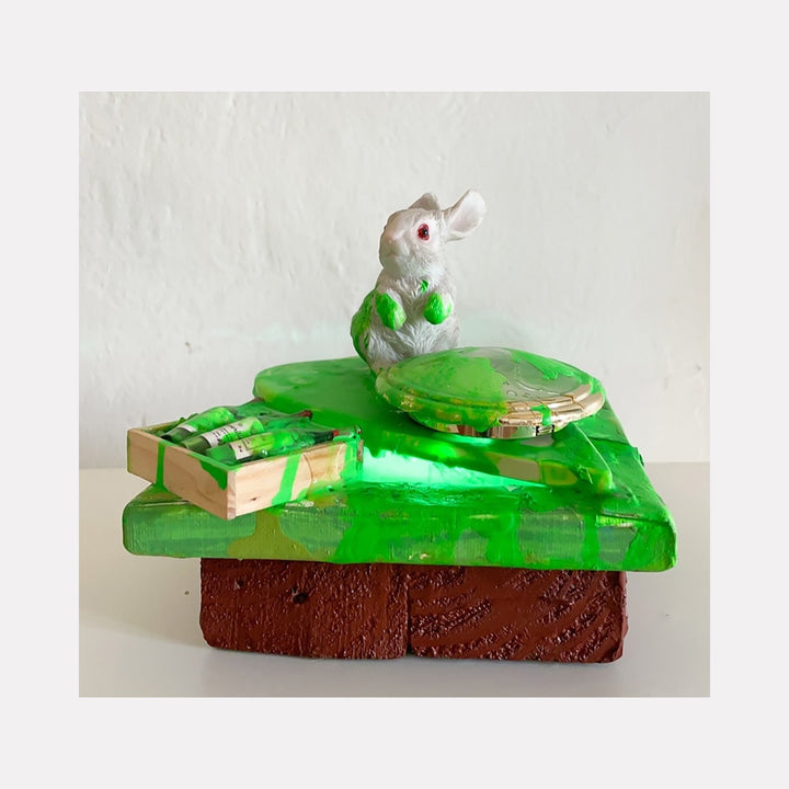 The artwork 'Bunny at Seance' by Brandon Hodges features a white ceramic rabbit perched atop a vibrant green acrylic platform with embedded LED lights. The piece incorporates miniature wine bottles and a Ouija board planchette, set on a textured brown base. The mixed-media assemblage glows with an ethereal green illumination.