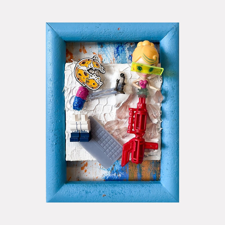 The artwork 'Digital Toybox' by Brandon Hodges features a vibrant blue wooden frame containing a mixed media assemblage with miniature toys, including a yellow toy head, red plastic components, and a metallic crown sticker against a textured white background with blue paint accents. 3D printed elements and plastic pieces create a playful, dimensional composition.
