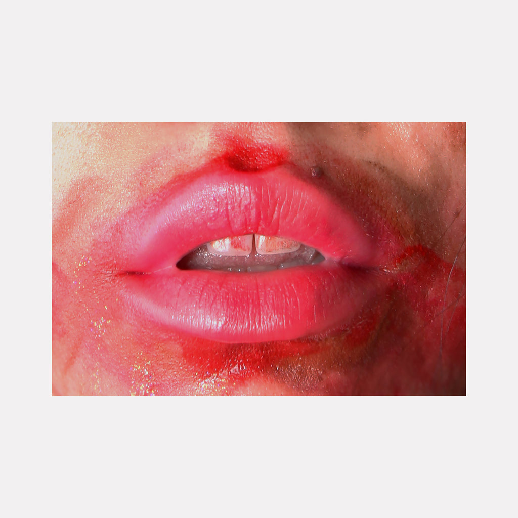 The artwork 'The Kissing Wall (After)' by Ronit Levin Delgado features a close-up photograph of vibrant red lips against a flesh-toned background. The glossy lips appear luminous with subtle textures and natural creases, surrounded by a soft red halo effect that diffuses into the skin. The artistic composition creates an intimate, abstract quality through its tight framing and rich color palette.