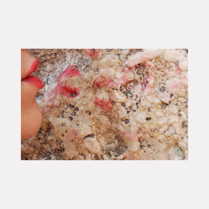 The artwork 'The Kissing Wall' by Ronit Levin Delgado captures a close-up photograph of a textured marble surface with pink and red lipstick marks scattered across its speckled beige and gray surface, creating an intimate abstract composition showing traces of human interaction. Limited edition glossy photographic print.