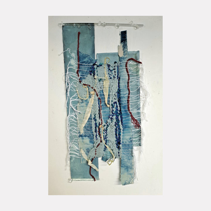 The artwork 'To The Skies' by Johanna Ryan features a vertical mixed-media composition in ethereal blue tones created through cyanotype process. Abstract woven elements combine canvas strips, natural twine, and dried corn husks, creating textural depth against geometric panels. The piece evokes a sense of upward movement through layered materials and frayed edges mounted on pale blue-tinted paper.